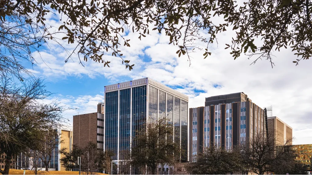Modern multi-story office buildings framed by tree branches under a partly cloudy blue sky.