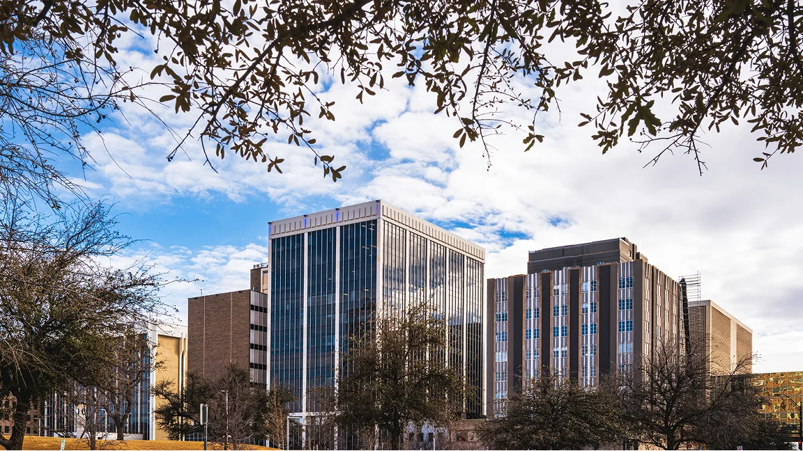 Modern multi-story office buildings framed by tree branches under a partly cloudy blue sky.