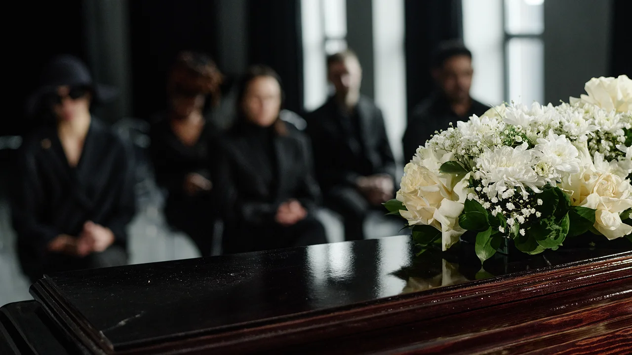 Mourners dressed in black sitting behind a wooden casket adorned with white flowers at a funeral.