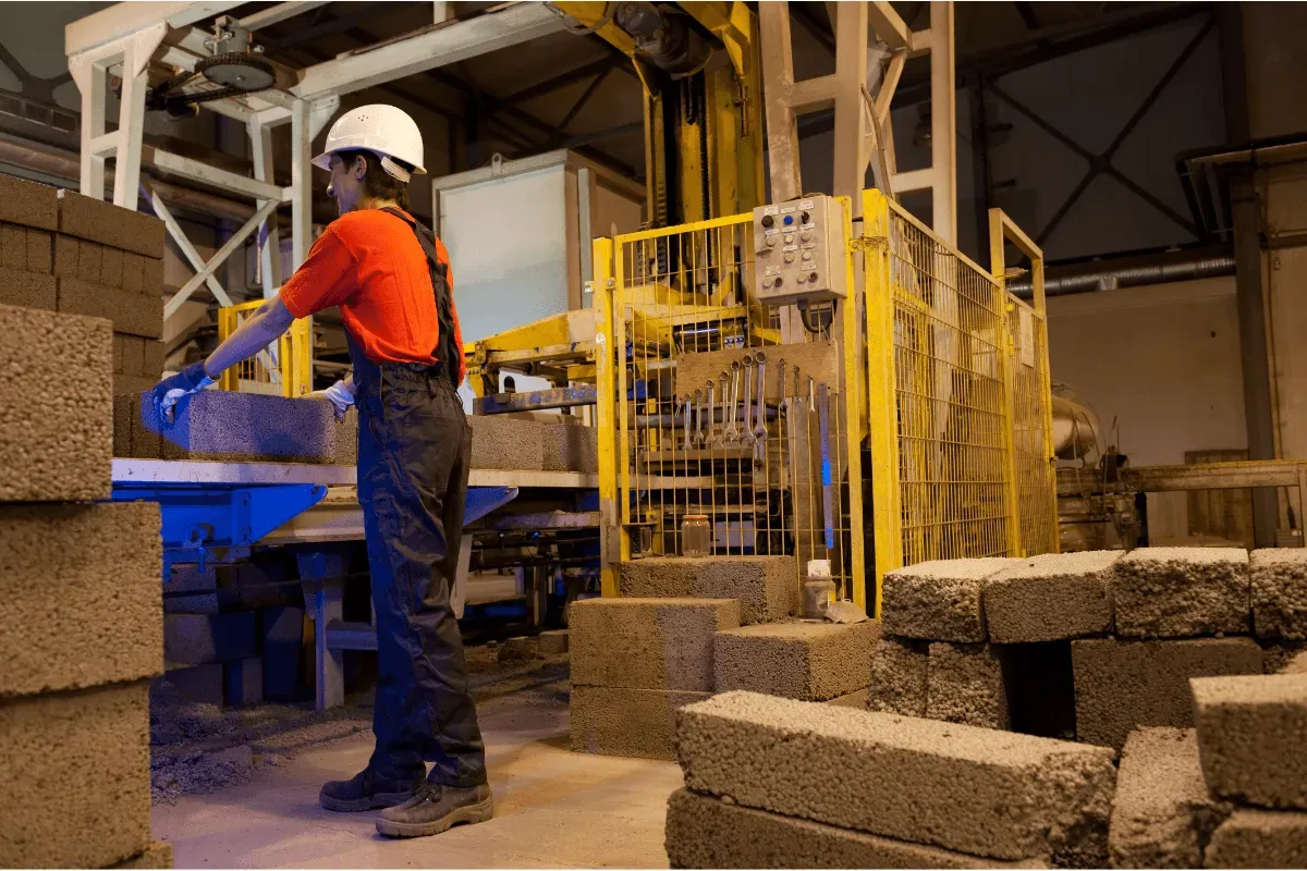 A factory worker in a hard hat and overalls moves concrete blocks on a production line.