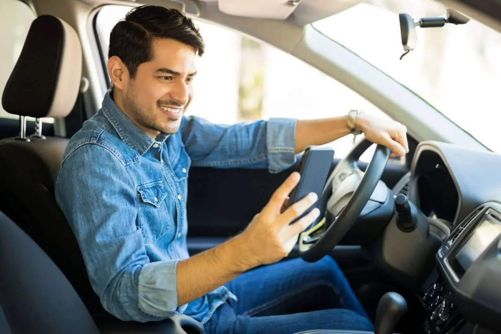 shows a smiling man sitting in the driver's seat of a car, looking at a smartphone in one hand while resting the other on the steering wheel.