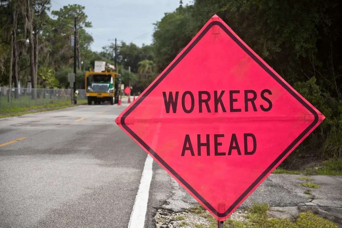 A bright orange-pink diamond road sign reading "WORKERS AHEAD" stands in the foreground, with a utility truck blurred in the background.