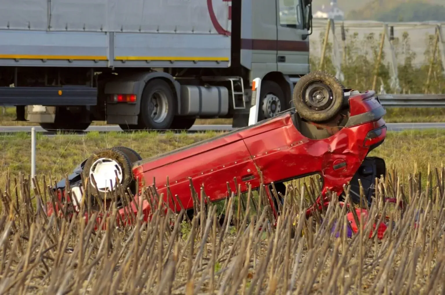 An overturned red car lying upside down in a field next to a highway, with a large truck driving past.