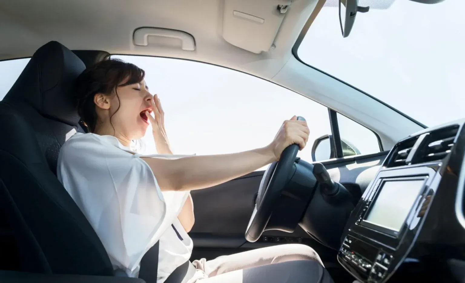 A tired woman yawning and covering her mouth with one hand while holding the steering wheel in the driver's seat of a car.