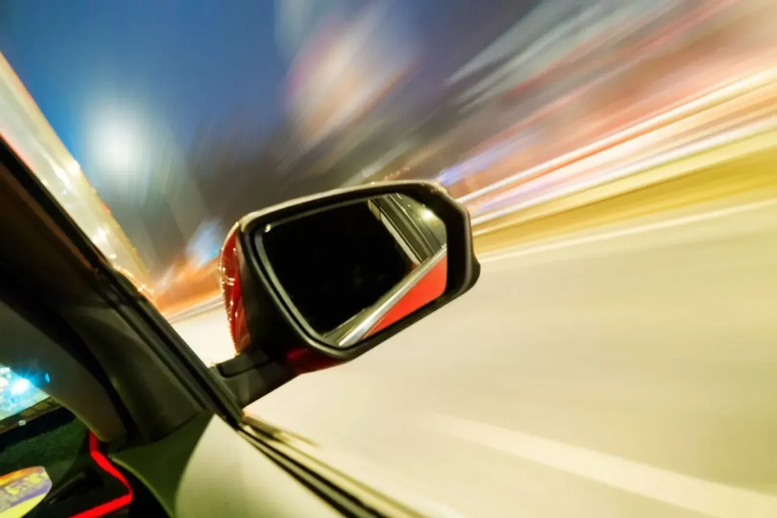 A close-up of a car's side mirror at night, surrounded by motion-blurred streaks of city lights.