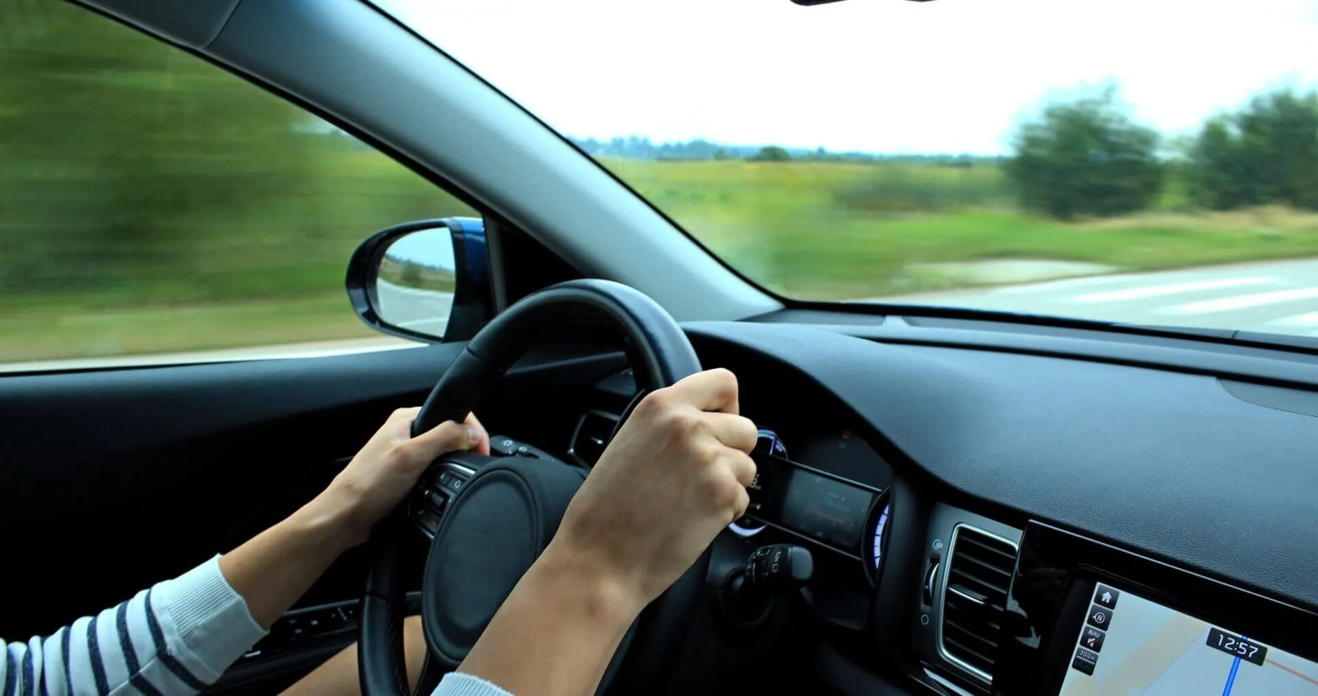 shows an interior view of a moving car, focusing on the driver's hands firmly gripping the steering wheel, with the scenery outside the window blurred from motion.