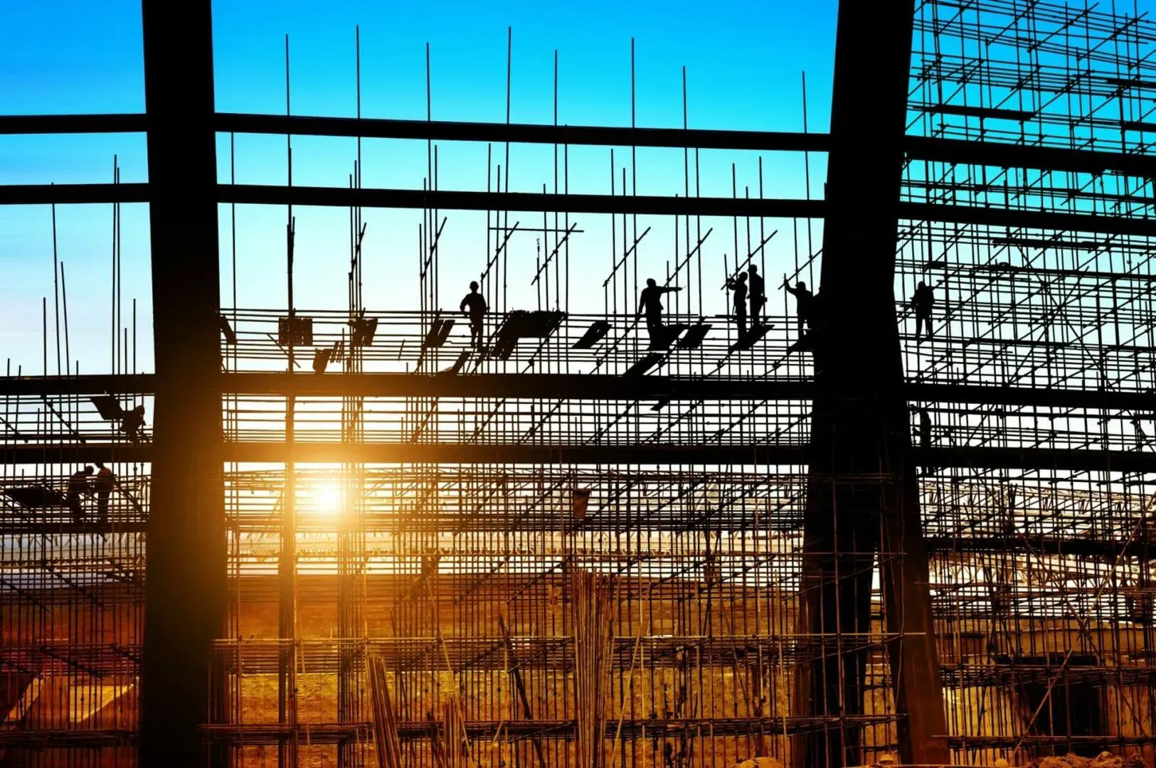 shows the silhouette of several construction workers standing on a large framework of scaffolding against a vibrant, setting sun.