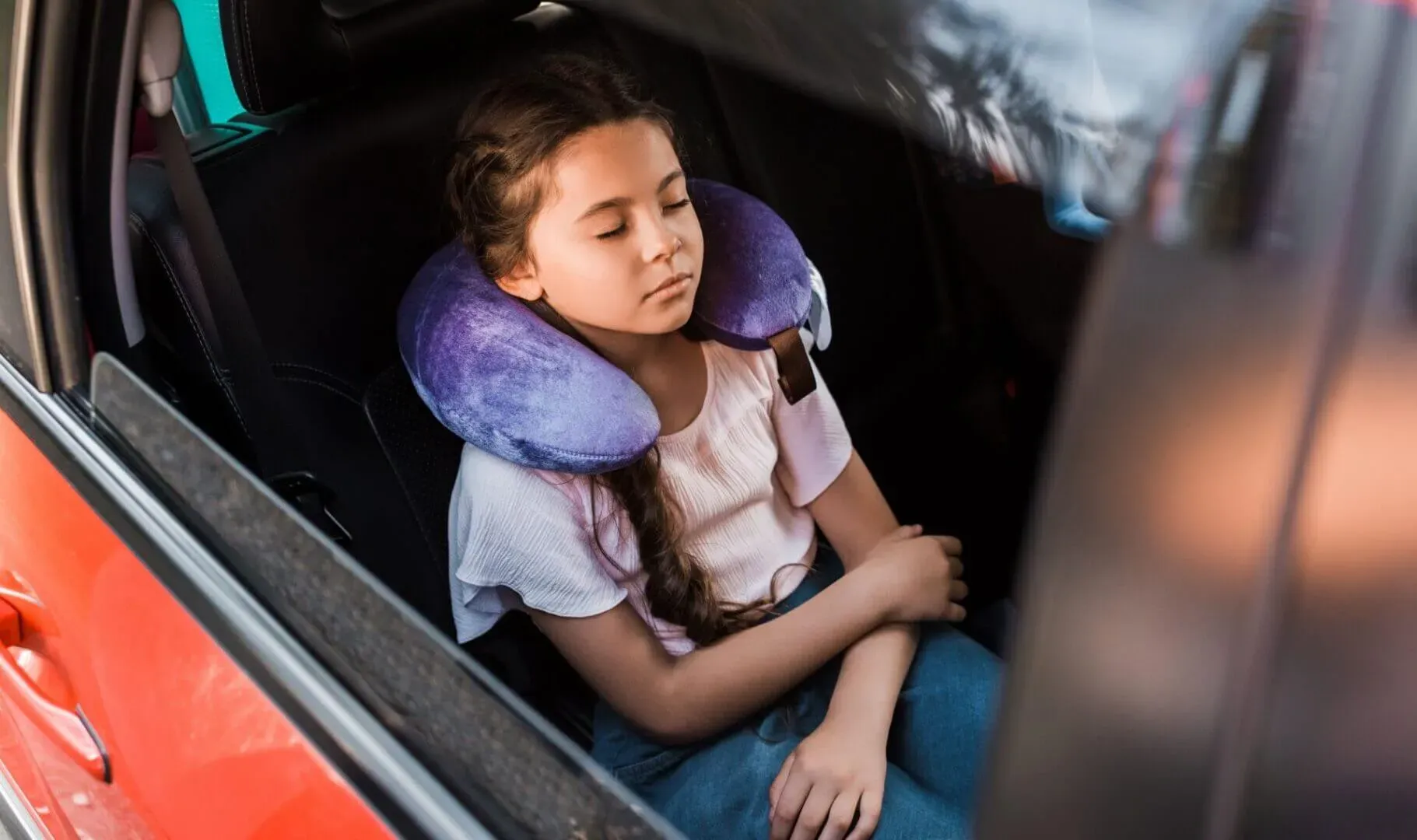 shows a young girl sleeping in the back seat of a car, wearing a purple neck pillow.