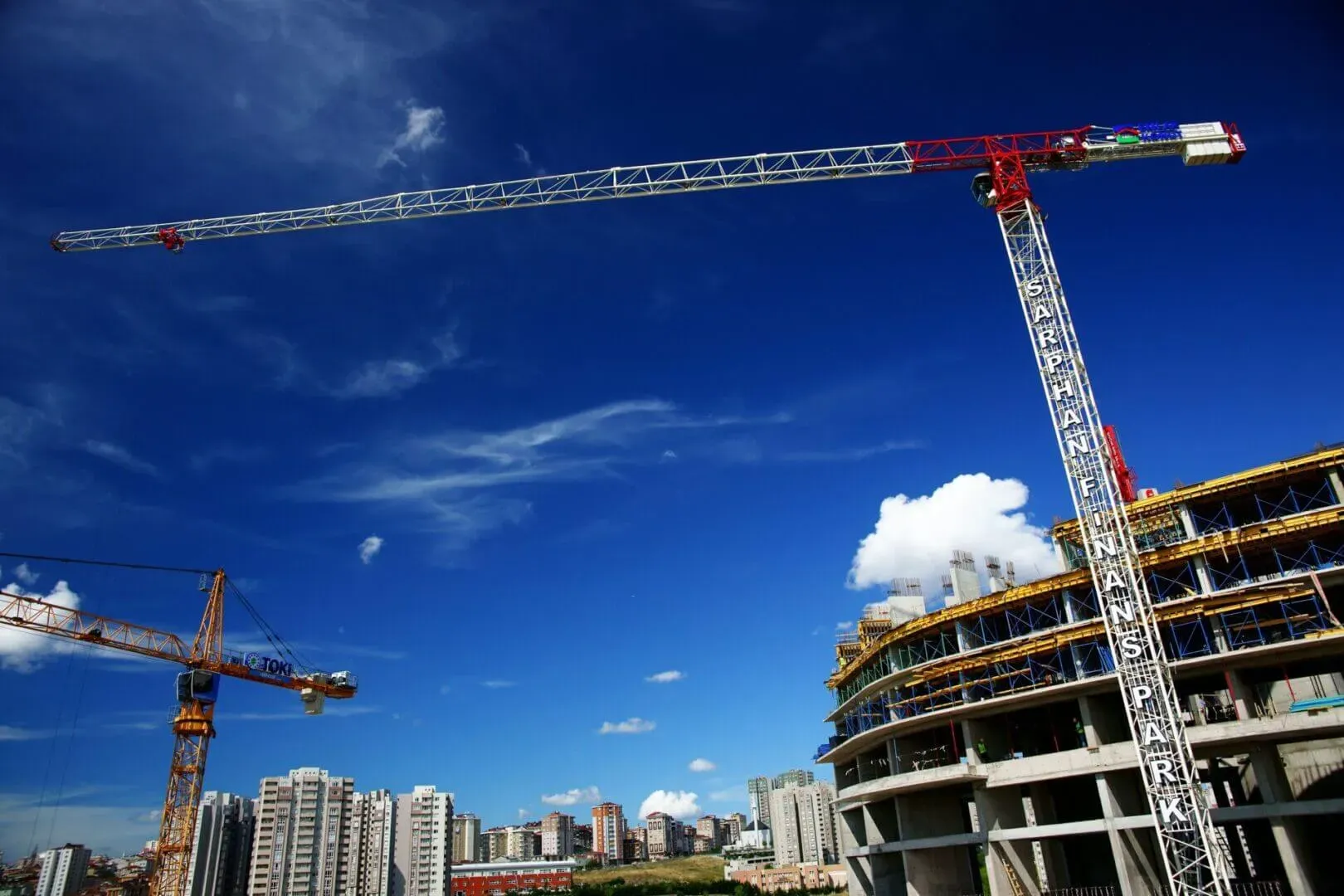 shows two tower cranes positioned over a large multi-story building under construction, set against a bright blue sky with a city skyline in the background.