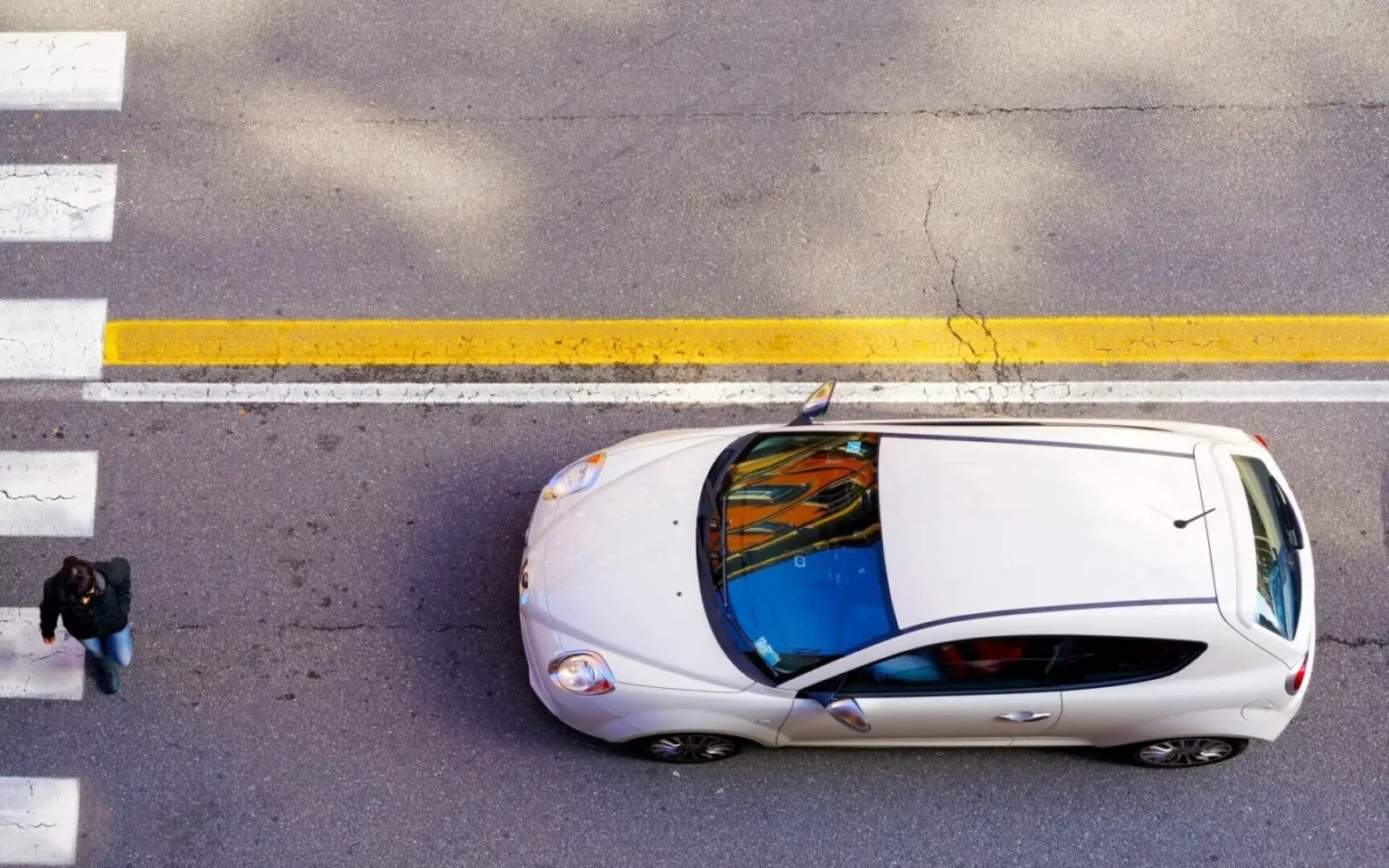 A white hatchback stops by a yellow line as a pedestrian crosses a crosswalk to the left.