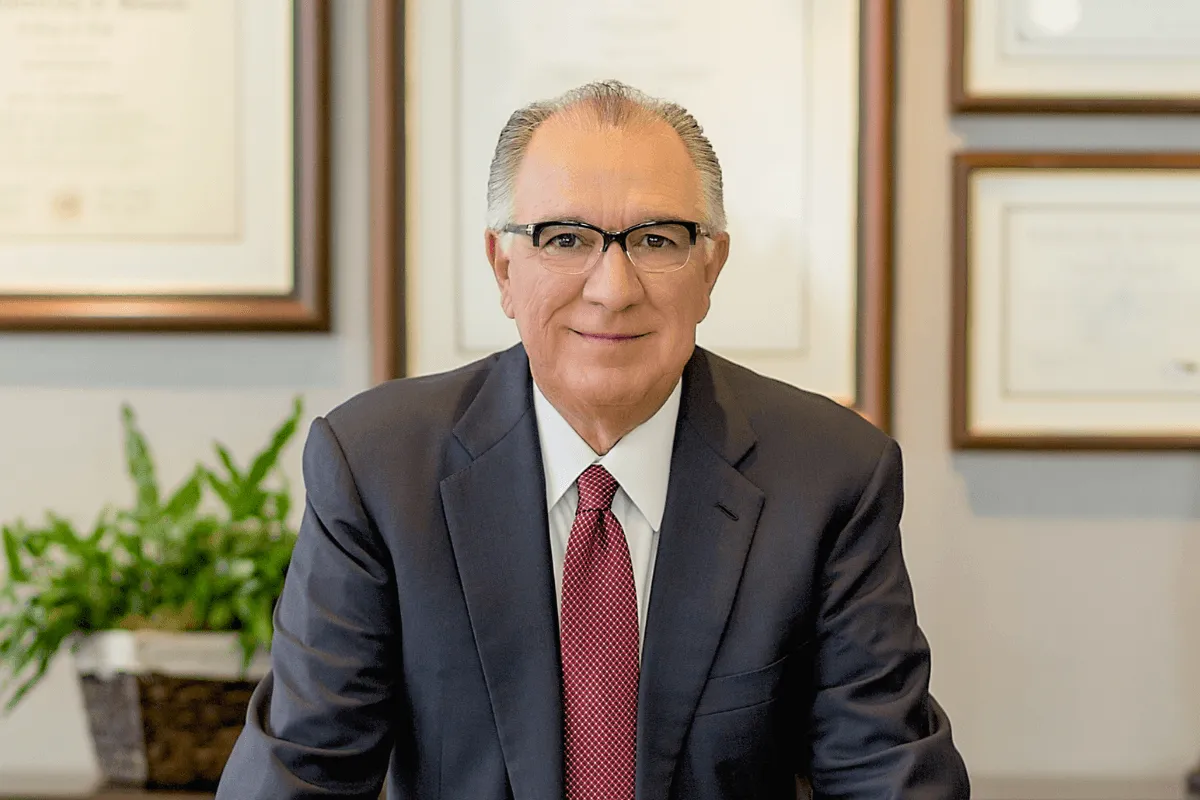 A professional portrait of a smiling man in a business suit, seated in an office with framed certificates in the background.