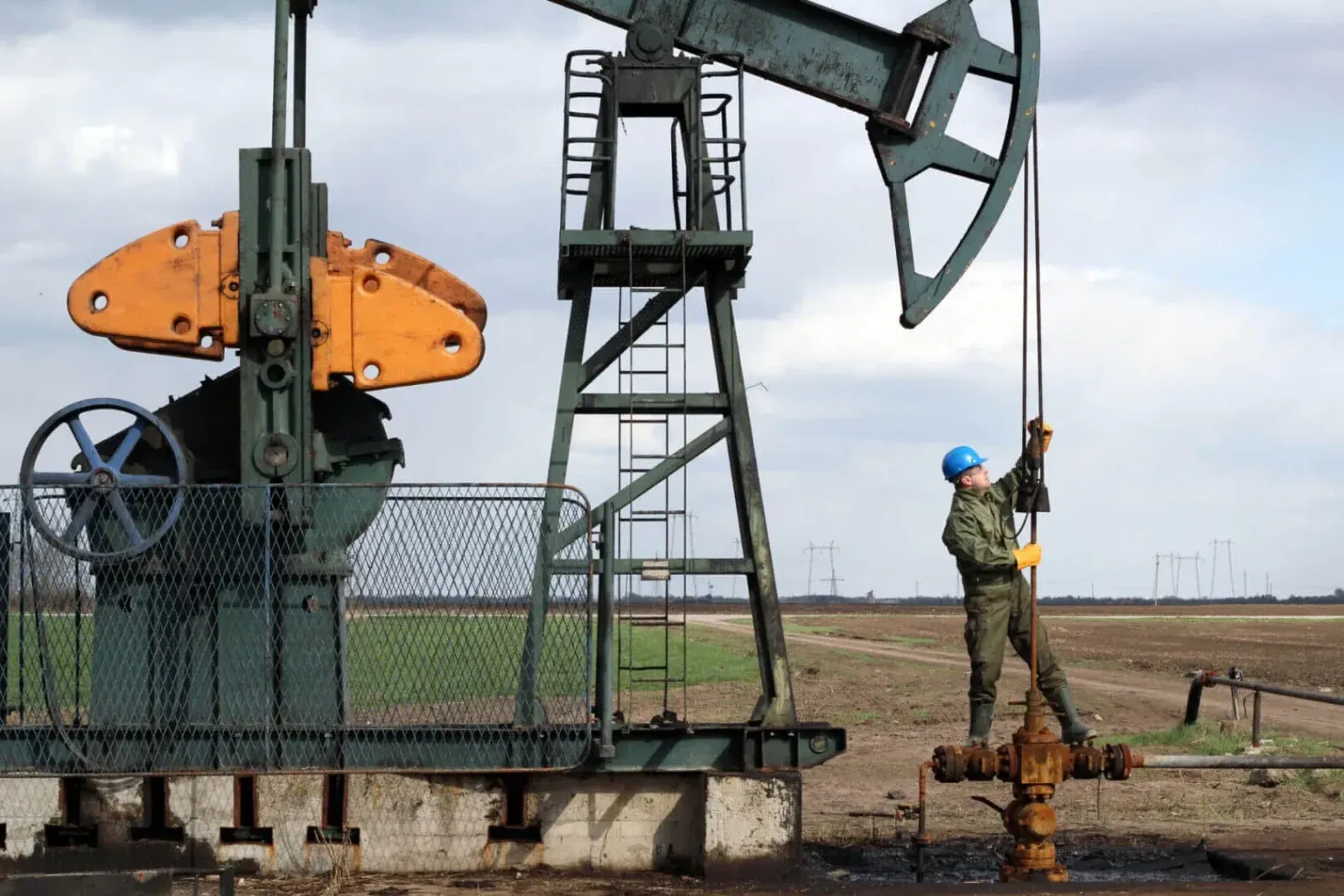 An oil field worker in a blue hard hat and green uniform maintains a pumpjack in an open field.