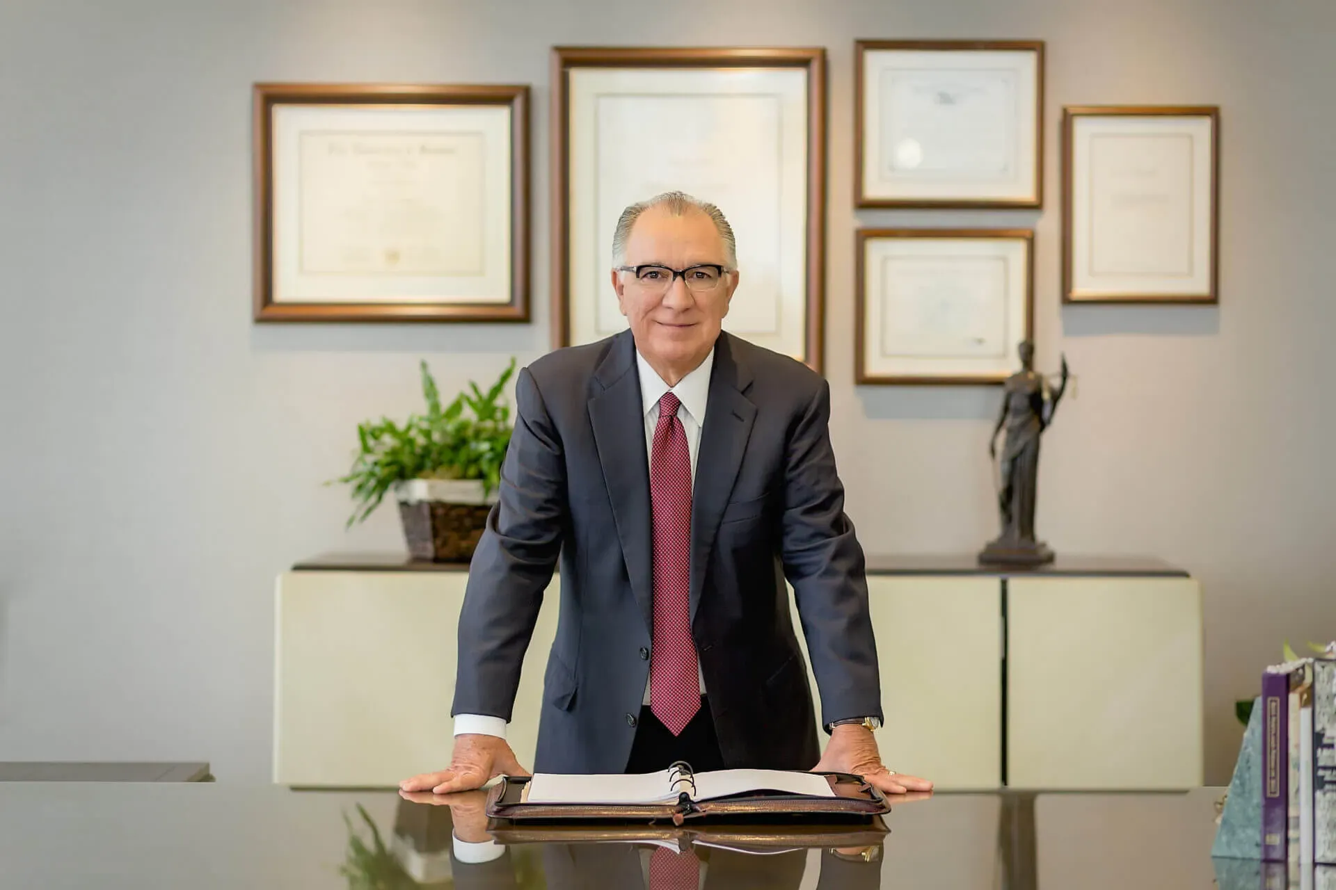 shows a man in a suit and tie leaning forward on a desk in a professional office setting, with framed certificates and a Lady Justice statue visible in the background.