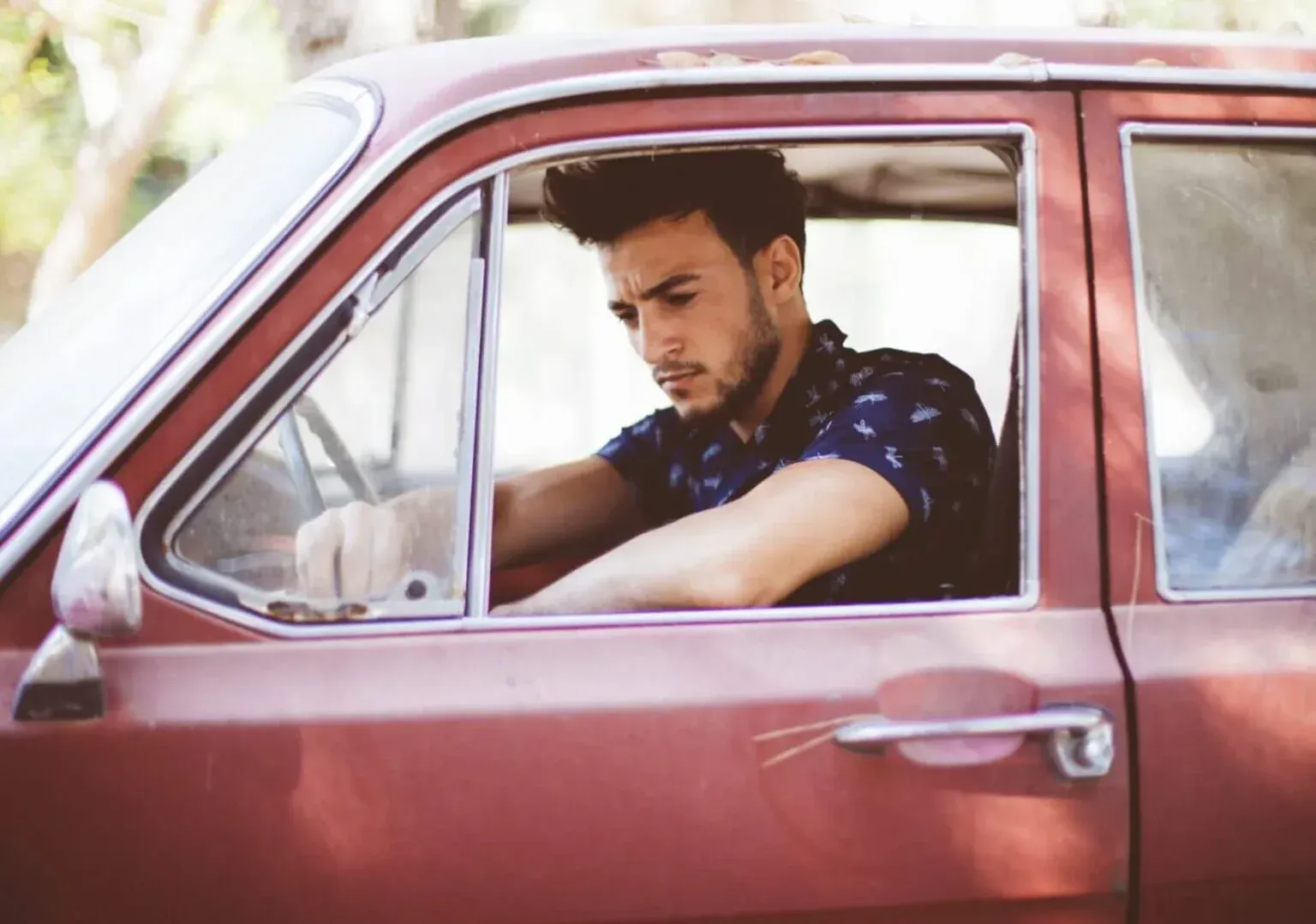 A young man looking down thoughtfully while sitting inside a vintage car, viewed through the open window.