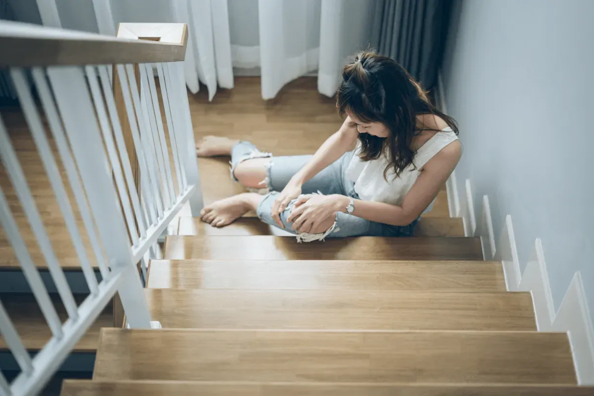 a woman sits injured on wooden stairs, holding her knee in pain after a slip and fall.