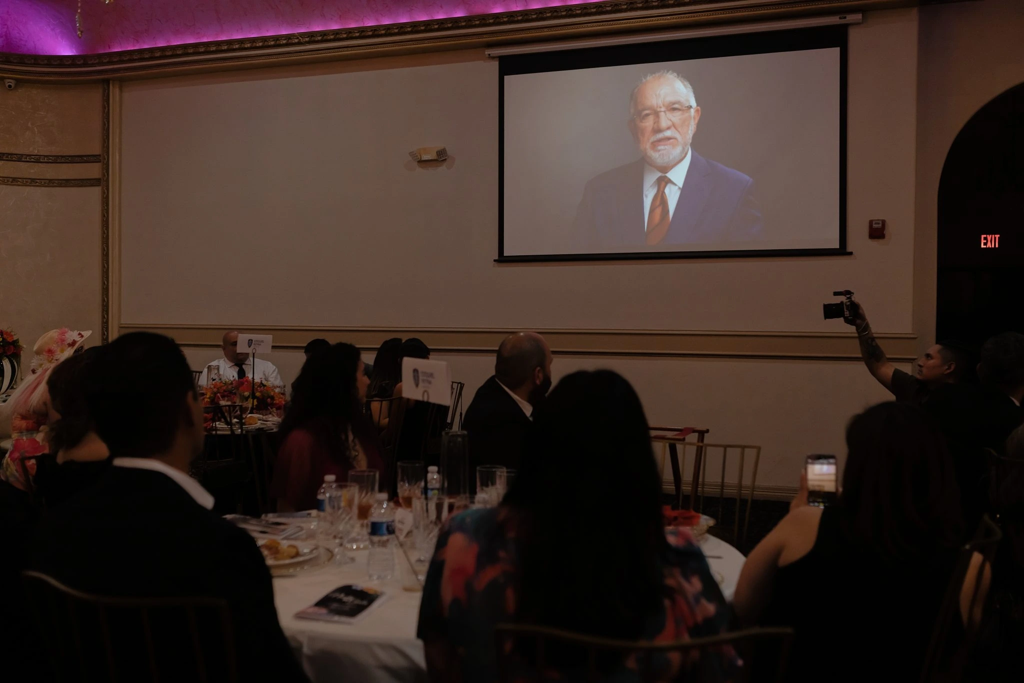 An audience at a banquet hall watches an older man in a suit speaking on a large projector screen.