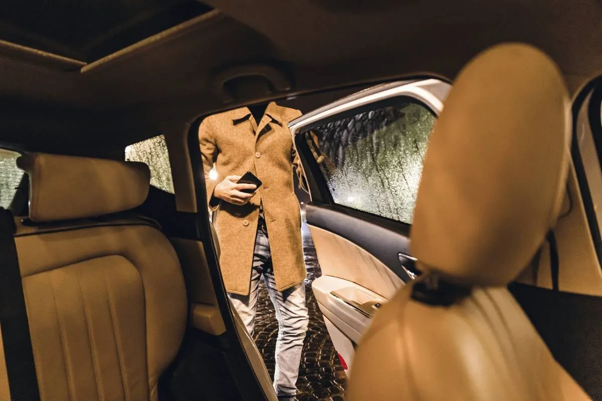 A man in a tan coat and jeans holding a smartphone steps out of a car at night on a rainy day.