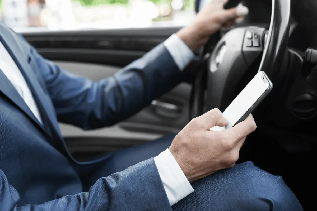 shows a person wearing a blue suit driving a car with one hand on the steering wheel while holding a smartphone in the other hand.