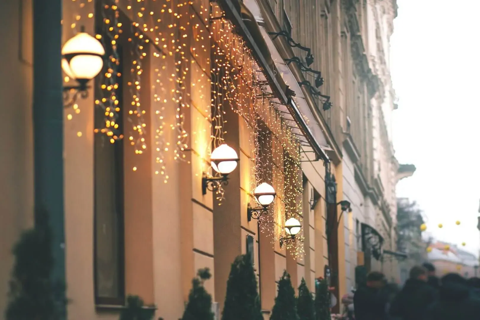 shows a building facade decorated with glowing, hanging string lights and round wall lamps, with a blurred crowd of pedestrians walking along the street in the background.