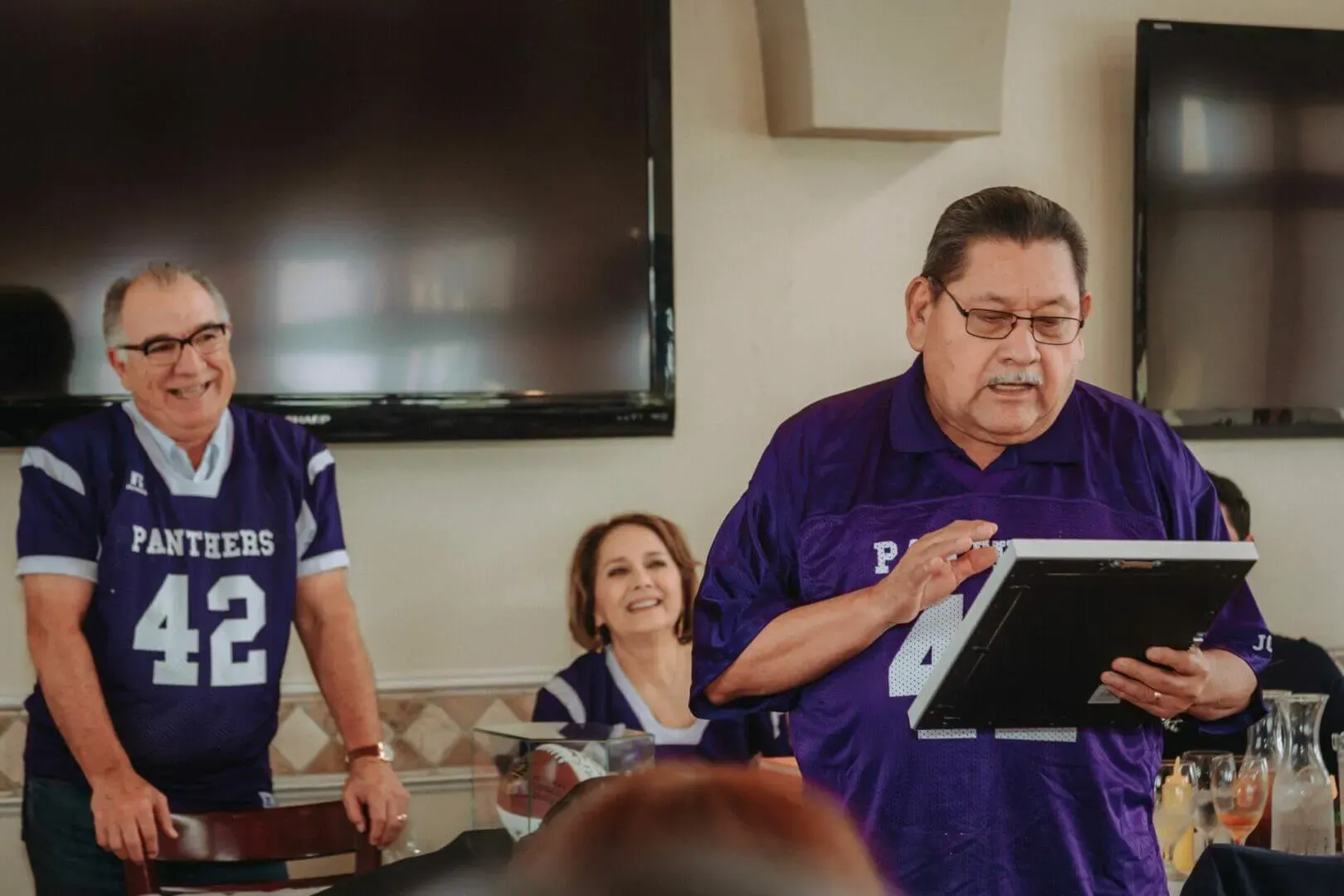 shows a small group of people at a gathering wearing purple "Panthers" football jerseys. One man is speaking and holding a framed object, while a man and a woman look on and smile.