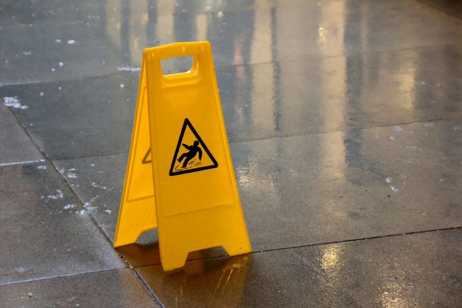 shows a yellow caution sign with a slipping figure icon placed on a wet, reflective tiled floor.