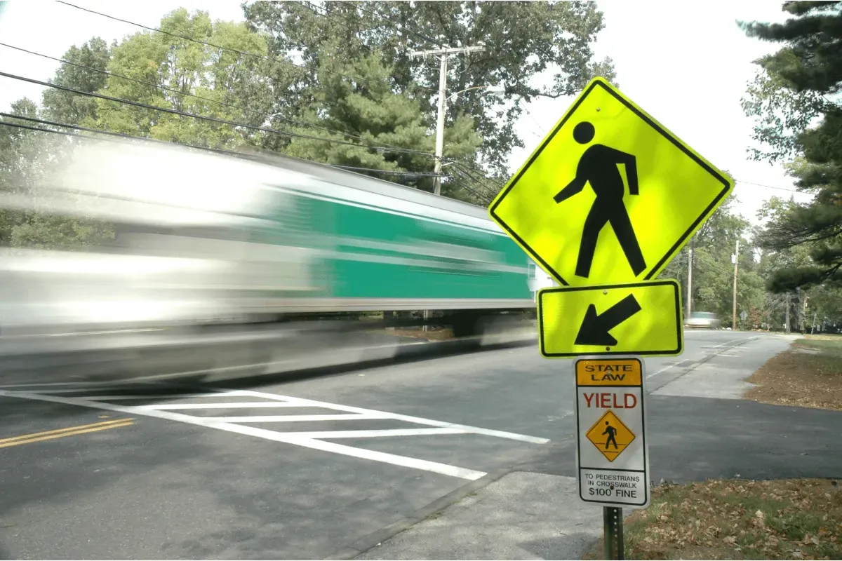 A semi-truck with a green and white trailer is captured in a motion blur as it drives past a marked pedestrian crosswalk. In the foreground on the right, a bright yellow-green diamond-shaped pedestrian crossing sign stands with a downward-pointing arrow and a smaller "State Law: Yield to Pedestrians" sign attached below it.
