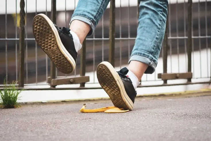 A person's foot is shown stepping onto a banana peel on the ground.