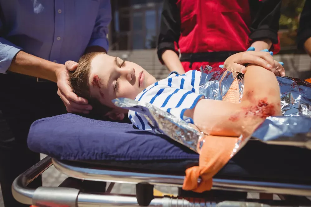 A young boy with visible injuries lies on a stretcher while being attended to by emergency personnel.