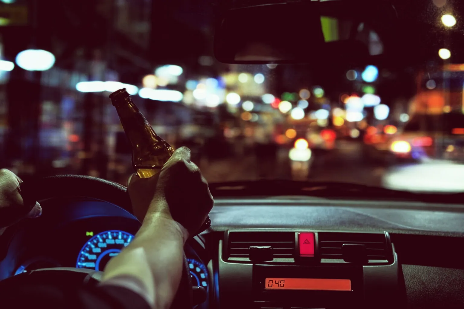 A person inside a car at night, holding a beer bottle while driving.