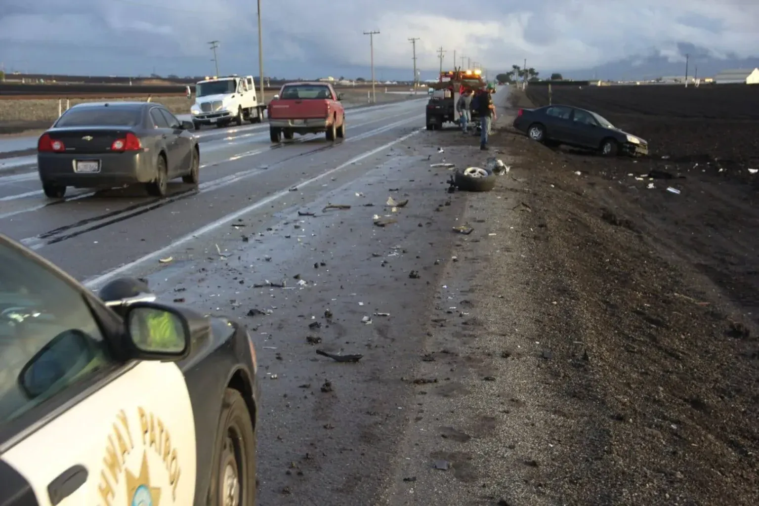A highway accident scene with debris scattered on the road, a car on the shoulder, and a Highway Patrol vehicle.