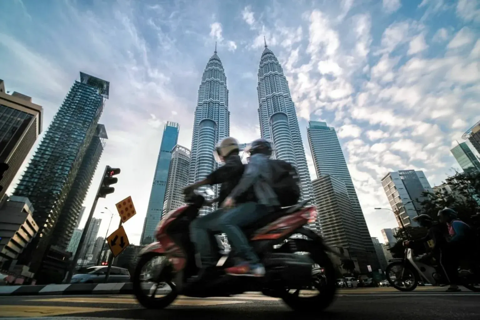 A scooter down a city street, with towering modern skyscrapers rising into a cloudy sky.