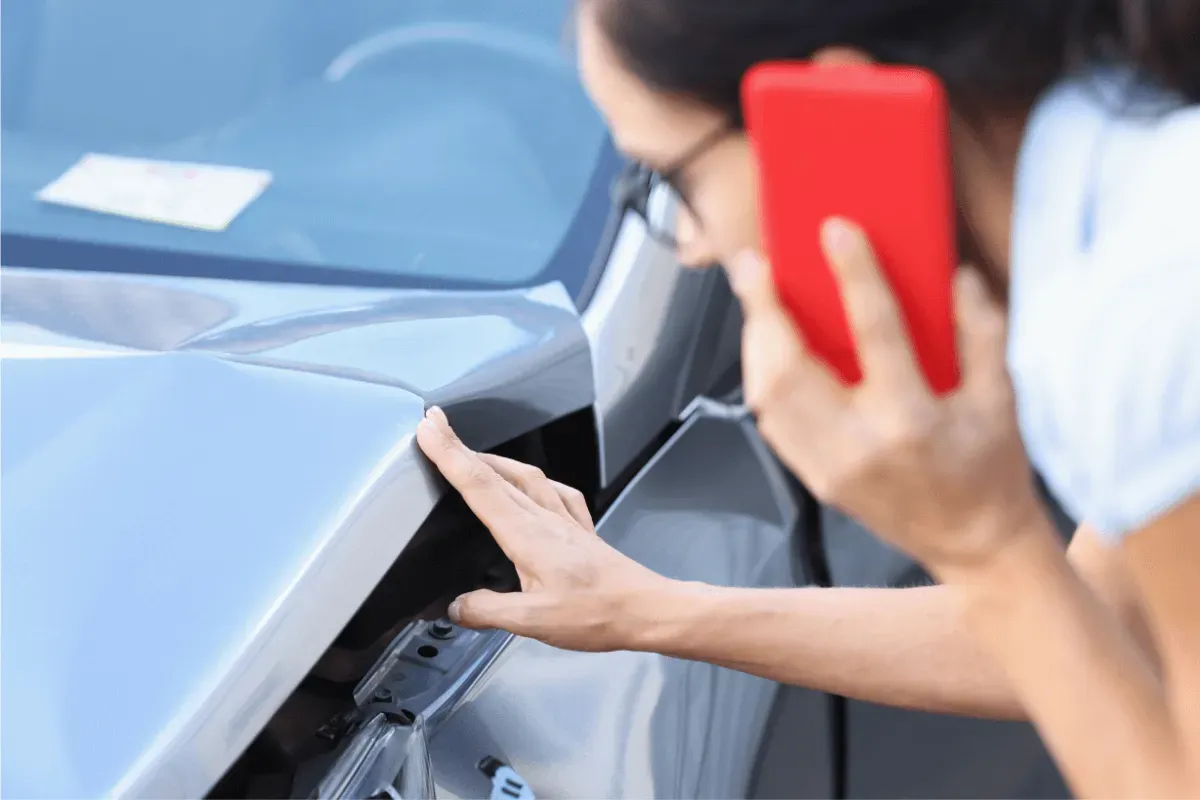 A person holding a red smartphone to their ear while inspecting a dented gray car hood after an accident.
